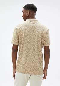 Camiseta polo de manga corta beige con un patrón floral. Presenta un cuello y un acabado texturizado, costuras visibles y un dobladillo ligeramente curvado.