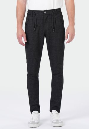 Mann trägt schwarz karierte Slim-Fit-Hose mit vorderen Reißverschlusstaschen und Kordelzug, kombiniert mit weißen Sneakern und weißem Hemd.