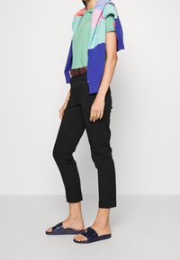 Avslappnad outfit med en mintgrön polo, svarta skräddarsydda byxor, en flerfärgad stickad tröja över axlarna och marinblå sandaler.