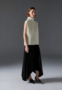 Mouwloze, geribbelde crèmekleurige coltrui gecombineerd met een zwarte geplisseerde rok met een asymmetrische zoom. Het model draagt zwarte puntige hakken.