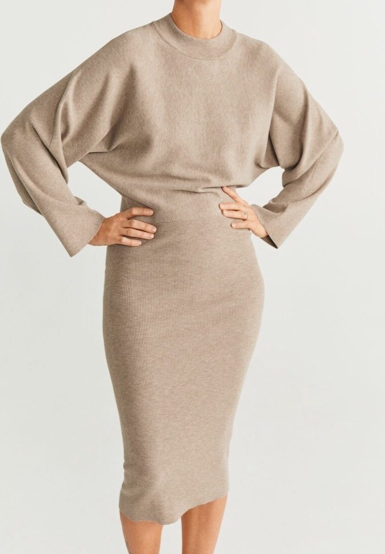 Femme portant un pull beige à manches longues et une jupe midi en maille côtelée assortie, les mains posées sur les hanches devant un fond uni.
