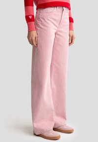 Světle růžové džíny s širokými nohavicemi z denimu, s vysokým pasem, bočními kapsami a jemnými šicími detaily. Kombinovány s světle růžovými teniskami.