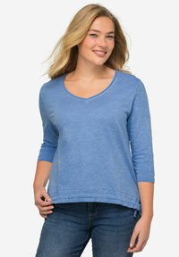 Top blu, a maniche lunghe, con scollatura a V e finitura texturizzata. Caratterizzato da un orlo curvo con nastri decorativi ai lati. Indossato con jeans di denim.
