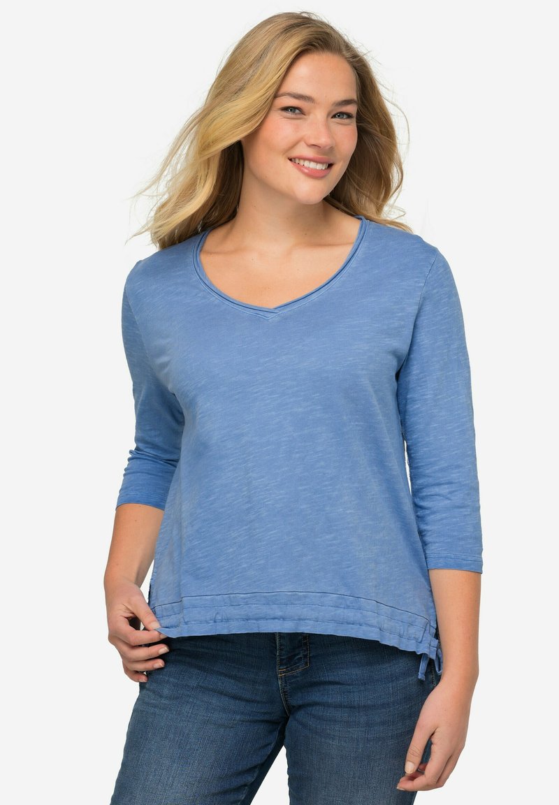 Top blu, a maniche lunghe, con scollatura a V e finitura texturizzata. Caratterizzato da un orlo curvo con nastri decorativi ai lati. Indossato con jeans di denim.