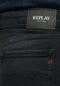 Πίσω τσέπη μαύρου τζιν με μαύρη δερμάτινη ετικέτα που γράφει "REPLAY SINCE 1981" και κόκκινο κεντητό "R" στη γωνία της τσέπης.