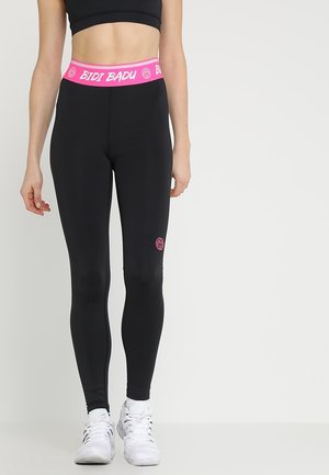 Vrouw draagt zwarte leggings met een roze tailleband met het label "BIDI BADU" en witte sportschoenen, staand tegen een effen achtergrond.