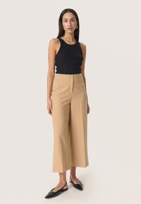 Top nero a costine, pantaloni beige a gamba larga con una texture liscia, dotati di chiusura frontale e tasche laterali, abbinati a mocassini neri.