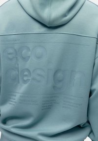 Heledsinine kapuutsiga sule, millel on tekstuuriga selja print ökokujunduse põhiprintsiipe, sealhulgas fraasid nagu "FUNKTSIONAALSUS" ja "KESTVUS".