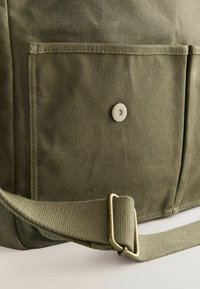 Canvas-Tasche in Olivgrün mit einer Vordertasche, Druckknopfschluss und verstellbarem Träger. Strukturierter Stoff mit sichtbaren Nähten und Metallbeschlägen.