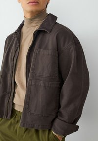 Brązowa bawełniana kurtka z dużymi kieszeniami z przodu, kołnierzem i luźnym krojem, połączona z beżowym prążkowanym golfem i zielonymi spodniami.