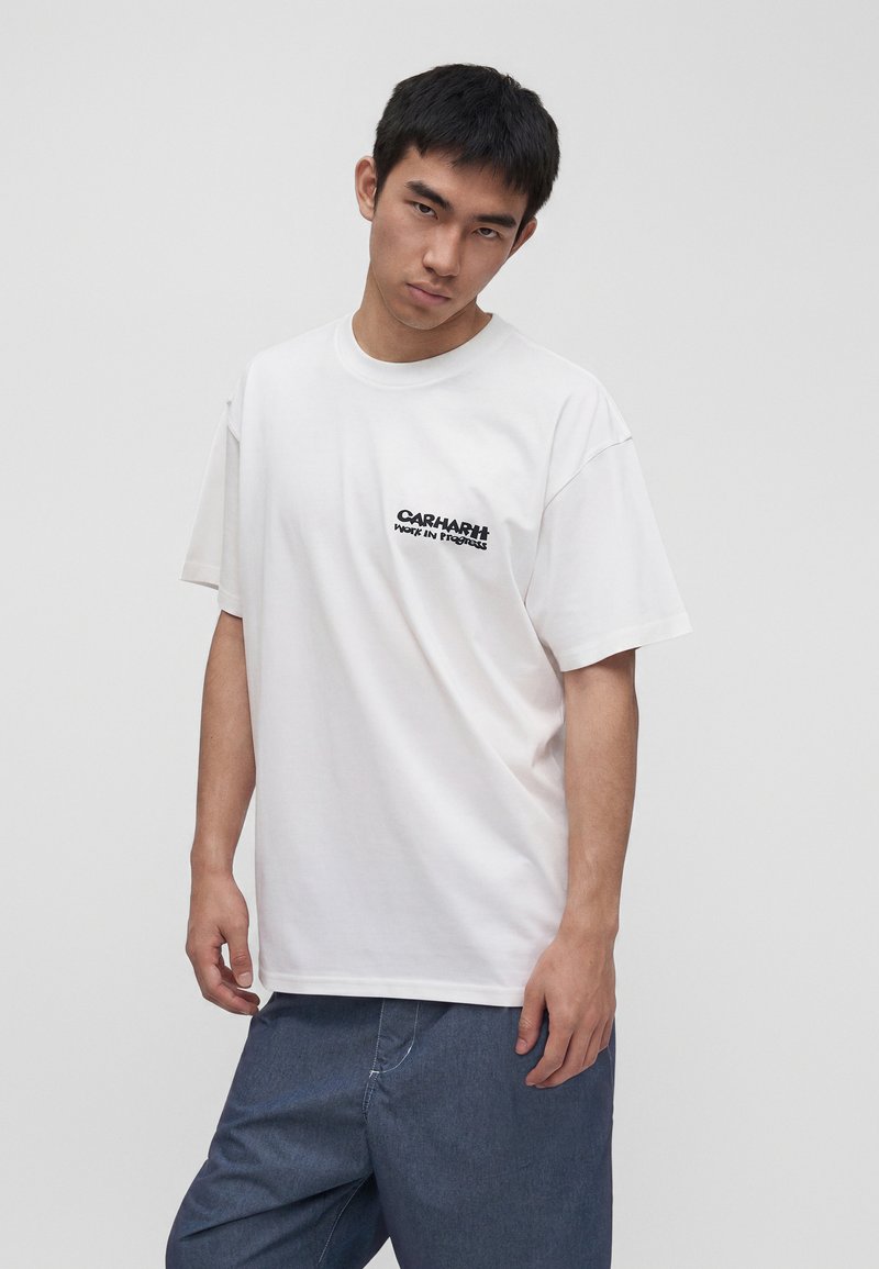 Giovane uomo che indossa una t-shirt bianca Carhartt con testo sul petto e pantaloni blu larghi, in piedi davanti a uno sfondo chiaro e semplice.
