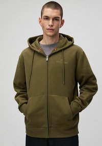 Oliwkowy hoodie na zamek z miękkiego materiału. Posiada kaptur ze sznurkiem, kieszenie z przodu i kontrastowy wykończenie wewnętrznego kaptura.