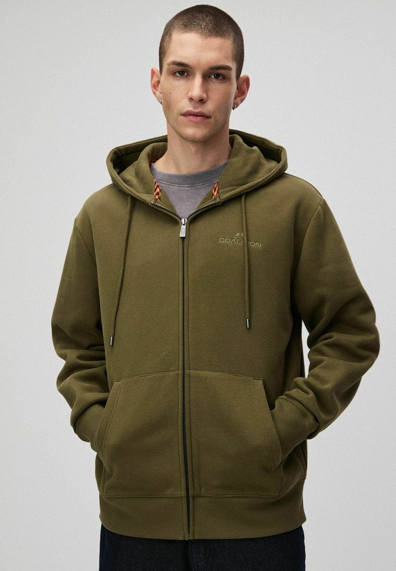 Oliwkowy hoodie na zamek z miękkiego materiału. Posiada kaptur ze sznurkiem, kieszenie z przodu i kontrastowy wykończenie wewnętrznego kaptura.