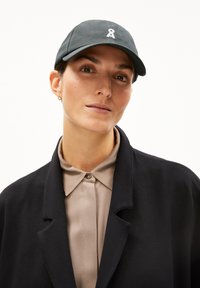 Jonge vrouw met een donkere pet met een wit embleem, beige overhemd met kraag dichtgeknoopt, en een zwarte jas tegen een effen achtergrond.