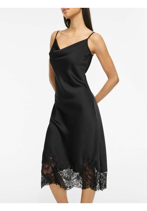 Femme portant une robe nuisette en satin noir avec fines bretelles et un ourlet orné de dentelle, debout les mains derrière le dos.