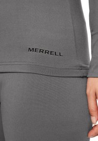 Szara, długi rękaw sportowa bluzka z płaskimi szwami i dopasowanym krojem. Posiada czarne logo "MERRELL" po lewej stronie, blisko dołu.