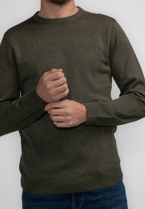 Mężczyzna noszący oliwkowo zielony dziany sweter z prążkowanymi mankietami i dołem, poprawiający rękaw, na jednolitym jasnym tle.