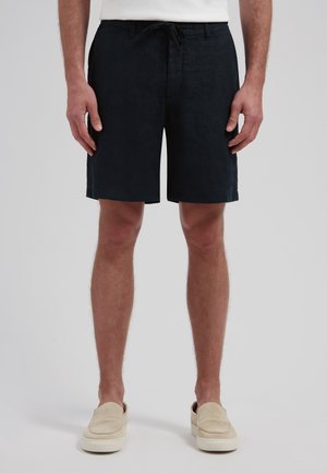 Man draagt zwarte shorts met trekkoord, beige instapschoenen en een wit shirt, staand tegen een effen grijze achtergrond.