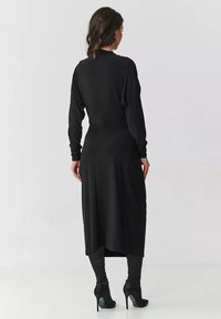 Czarna sukienka z długim rękawem i wysokim kołnierzem, z plisowanym tyłem i prostą sylwetką, zestawiona z czarnymi botkami na wysokim obcasie.