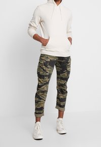 Kremowa bluza z kapturem z przednią kieszenią i sznurkiem, zestawiona z zielonymi moro spodniami cargo z wieloma kieszeniami i podwiniętymi nogawkami. Białe trampki.