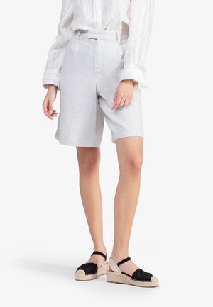 Femme portant un short gris clair sur mesure jusqu’aux genoux, une chemise blanche à manches longues avec des rayures verticales et des sandales compensées espadrilles noires.