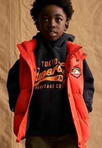 Gilet rouge rembourré sur un sweat à capuche bleu marine avec du texte orange. Le gilet a des boutons visibles et une patch sur l'épaule.