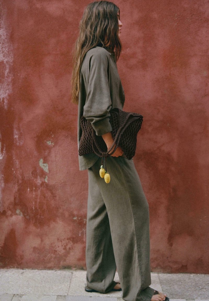 Femme en tenue ample vert olive tenant un grand sac tissé marron foncé, debout de profil contre un mur texturé rougeâtre.