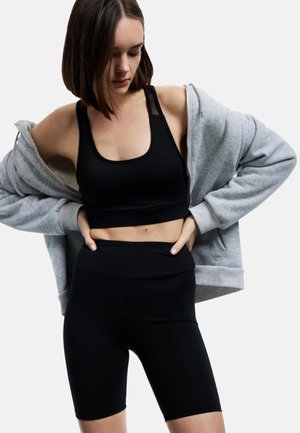 Frau trägt schwarzen Sport-BH und Radlerhose, zieht einen grauen Kapuzenpullover mit Reißverschluss an, schaut nach unten, isoliert auf weißem Hintergrund.