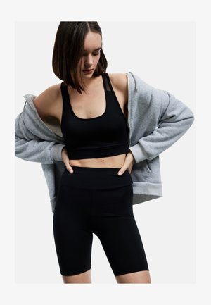 Frau trägt schwarzen Sport-BH und Radlerhose, zieht einen grauen Kapuzenpullover mit Reißverschluss an, schaut nach unten, isoliert auf weißem Hintergrund.