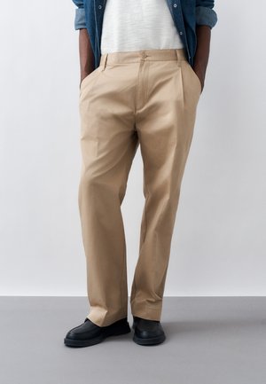 Mężczyzna w beżowych plisowanych spodniach, czarnych butach, białej koszuli i niebieskiej dżinsowej kurtce, stojący na szarej podłodze z rękami w kieszeniach.