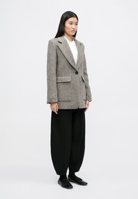 Γκρι σακάκι με καρώ σχέδιο και notch πέτοι, δύο μπροστινές τσέπες και κλείσιμο με έναν μόνο κουμπί, συνδυασμένο με χαλαρές μαύρες παντελόνες.