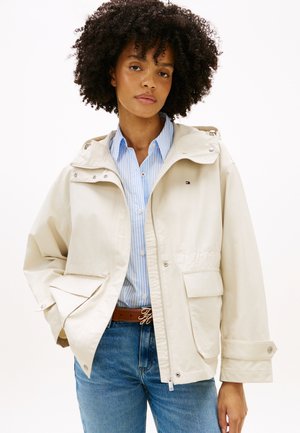 Kobieta o kręconych włosach, ubrana w beżową kurtkę z kapturem, na niebieskiej koszuli w paski zapinanej na guziki i niebieskich dżinsach, stojąca z jedną ręką w kieszeni.