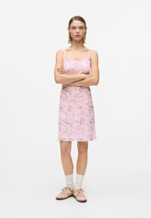 Ung kvinne som står med armene i kors iført en rosa blomstrete kjole, hvite sokker og rosa joggesko på en helt hvit bakgrunn.