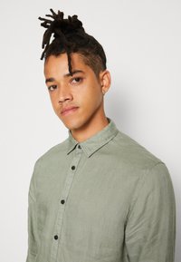 Camisa verde claro de botones con un acabado texturizado, que presenta un cuello puntiagudo y botones oscuros, llevada por una persona con el cabello corto.
