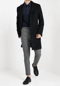 Czarny długi płaszcz na ciemnej koszuli, w połączeniu z szarymi obcisłymi spodniami i czarnymi tenisówkami. Charakteryzuje się gładką fakturą i dopasowanym krojem.