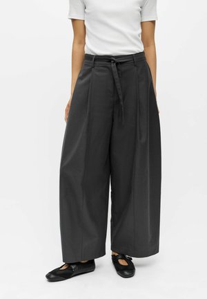 Femme portant un pantalon noir à jambes larges avec une ceinture nouée, une chemise blanche à manches courtes et des chaussures plates noires sur fond blanc.
