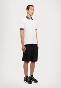 Muž v bielom polo tričku s krátkym rukávom a čiernym vzorovaným golierom a rukávmi, v čiernych voľných šortkách a čiernych teniskách s bielymi šnúrkami.