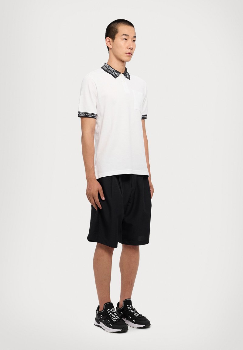 Muž v bielom polo tričku s krátkym rukávom a čiernym vzorovaným golierom a rukávmi, v čiernych voľných šortkách a čiernych teniskách s bielymi šnúrkami.