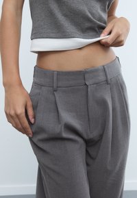 Graue, maßgeschneiderte Hose mit Falten, aus einem glatten Stoff und mit einem taillierten Bund. Hand auf der Taille positioniert. Weißes Oberteil ist darunter sichtbar.