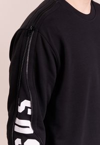 Sweat-shirt noir avec un détail de fermeture éclair sur l'épaule, présentant un texte blanc sur la manche. Confectionné dans un tissu doux avec une coupe ajustée.