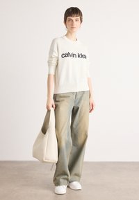 Kremfarget strikkegenser med svart "calvin klein" logo, løse blå jeans, hvite joggesko og en beige hobo-veske, stående mot en lys bakgrunn.