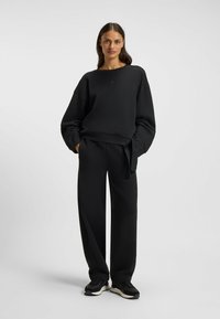 Svart oversized sweatshirt med ribbade ärmslut och en logotyp, tillsammans med högmidjade svarta byxor med knytning i midjan. Svarta sneakers.