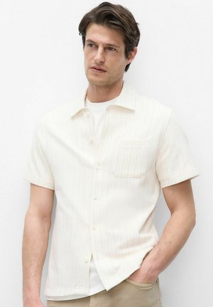 Mann mit braunen Haaren, der ein cremefarbenes kurzärmeliges Hemd über einem weißen T-Shirt trägt, mit einer Hand in der Tasche der beigen Hose.