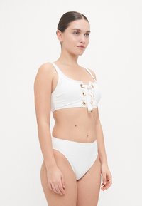 White bikini set featuring a cropped top with gold-tone eyelets and lace-up detailing, paired with high-waisted bottoms. Smooth, stretchy material.
