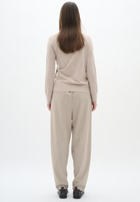 Beige Pullover mit einem taillierten Saum, kombiniert mit locker sitzenden, hellbeigen Hosen. Dunkles Schuhwerk rundet das Outfit ab. Schlichter Hintergrund.