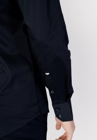 Maglietta a maniche lunghe navy con polsini abbottonati, orlo curvo e trama di tessuto morbida. Include dettaglio del polsino arrotondato con bottone a contrasto.