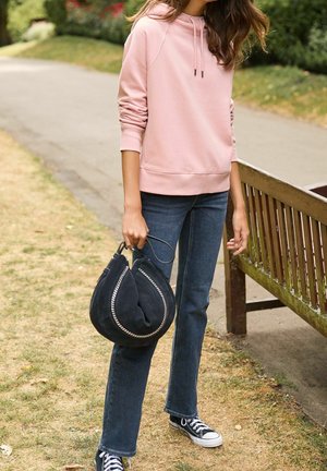 Lyserød hoodie med snore, blå denim flared jeans, sort taske med sølv hardware og sorte sneakers. Græs og stibaggrund.