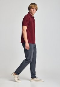Camisa polo burdeos de manga corta, combinada con pantalones gris oscuro. El modelo está de lado, llevando zapatillas crema, mostrando un look casual.