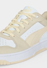 Gros plan d'une basket Puma beige et blanche avec des lacets crème, montrant le côté, le dessus et une partie de la semelle.