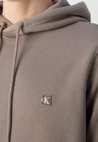 Gros plan sur un sweat à capuche taupe avec cordons de serrage et petite étiquette logo Calvin Klein sur la poitrine.